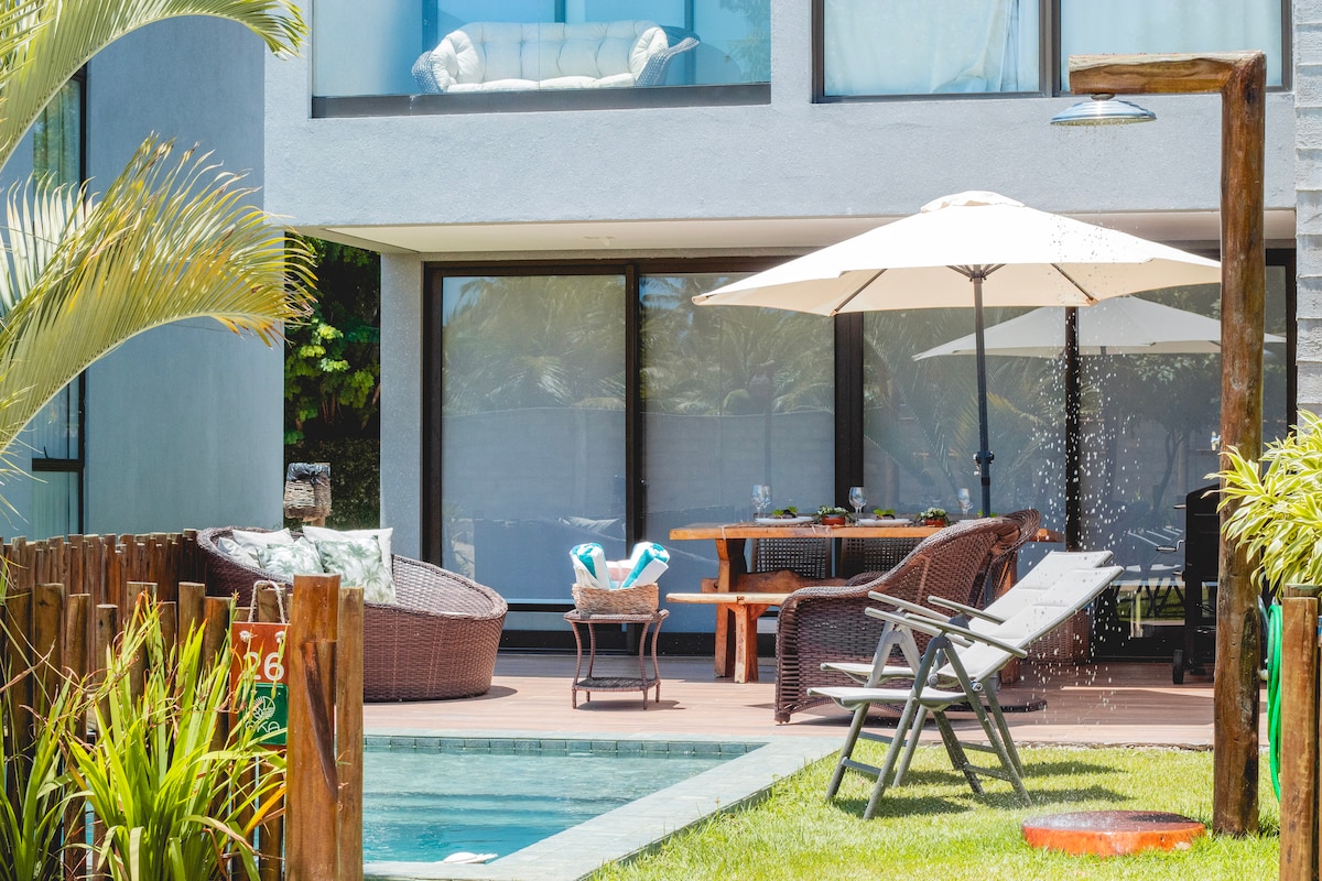 A private outdoor area is featured, complete with a swimming pool and comfortable seating, including woven lounge chairs and a dining table. An umbrella provides shade over the seating area, while tropical greenery and a wooden fence frame the space, enhancing its inviting atmosphere.