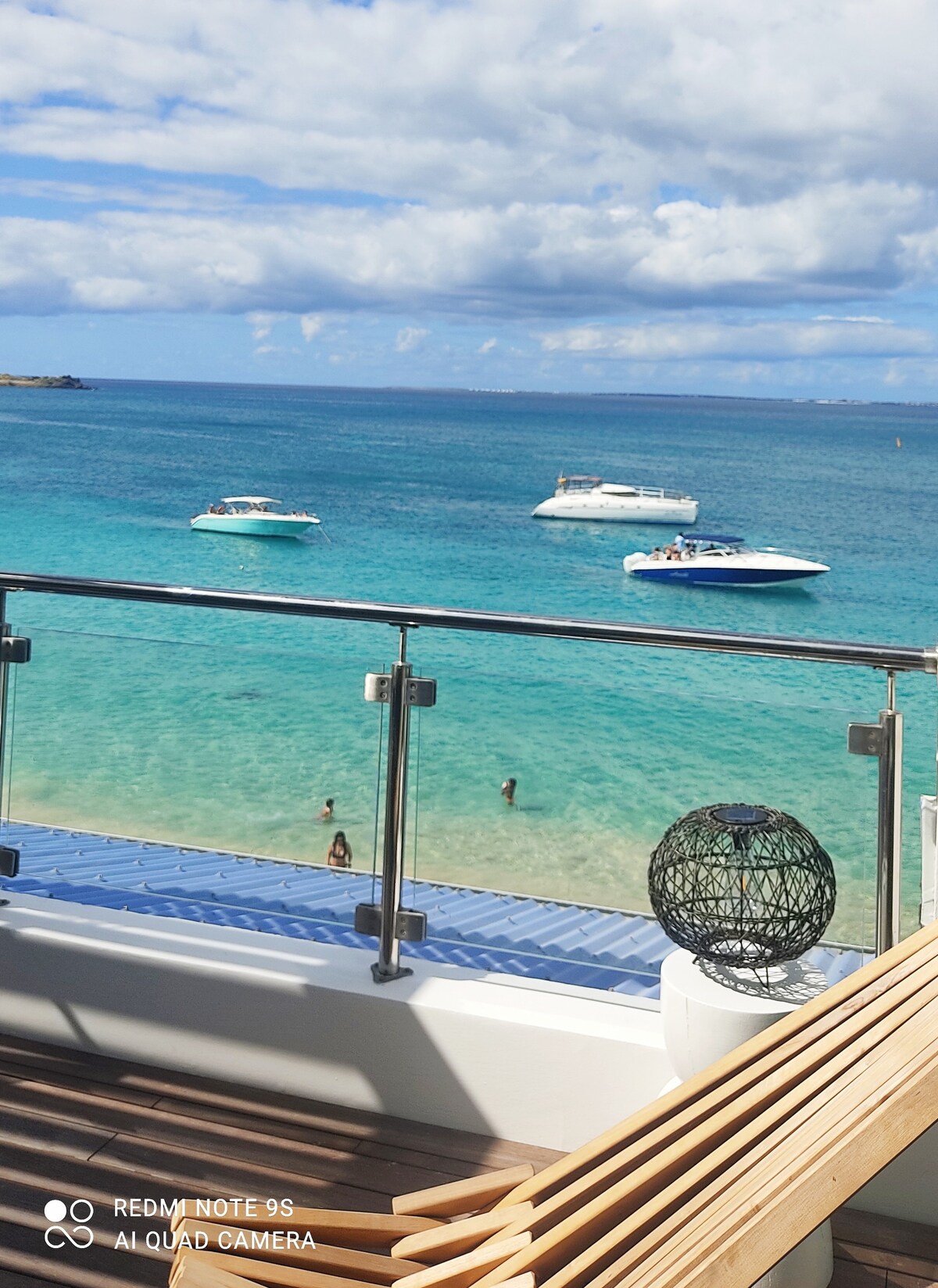 A view of crystal-clear Caribbean waters is captured from a balcony, where several boats float gently on the surface. The shoreline is visible in the distance, with a few people enjoying the beach. Sunlight reflects off the water, enhancing the tranquil scene.