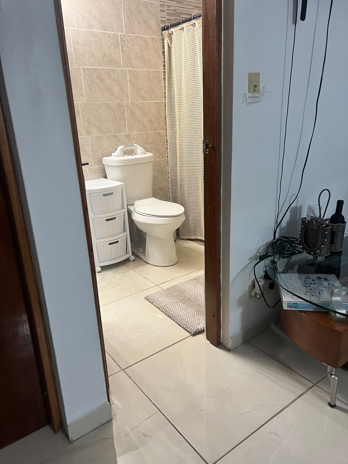 An accessible bathroom is visible, featuring neutral tiles and a compact layout. A toilet and a white storage unit are positioned on one side. Natural light is suggested through a curtain in the background, while the entryway offers a glimpse of the adjoining room.