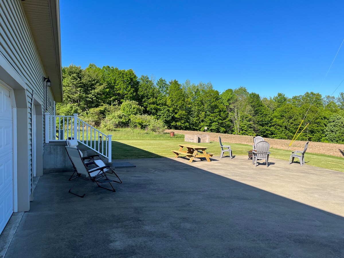 A spacious outdoor area features a concrete patio with a pair of lounge chairs, a picnic table, and several Adirondack chairs. Lush greenery surrounds the yard, offering a natural backdrop. The clear blue sky complements the tranquil setting, ideal for relaxation.