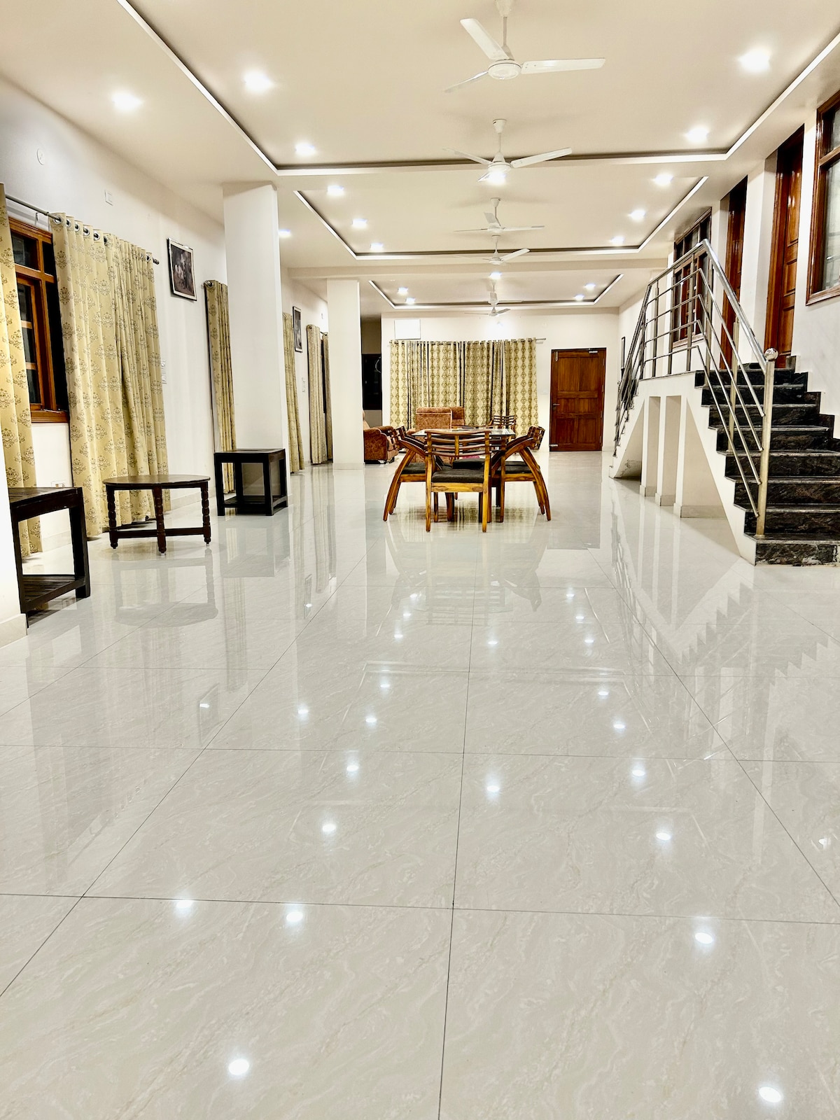 A spacious living and dining area is showcased, featuring a polished floor that reflects the overhead lights. A large wooden dining table is set near the windows, with ample space surrounding it. The room is illuminated by natural light and has multiple ceiling fans for airflow.