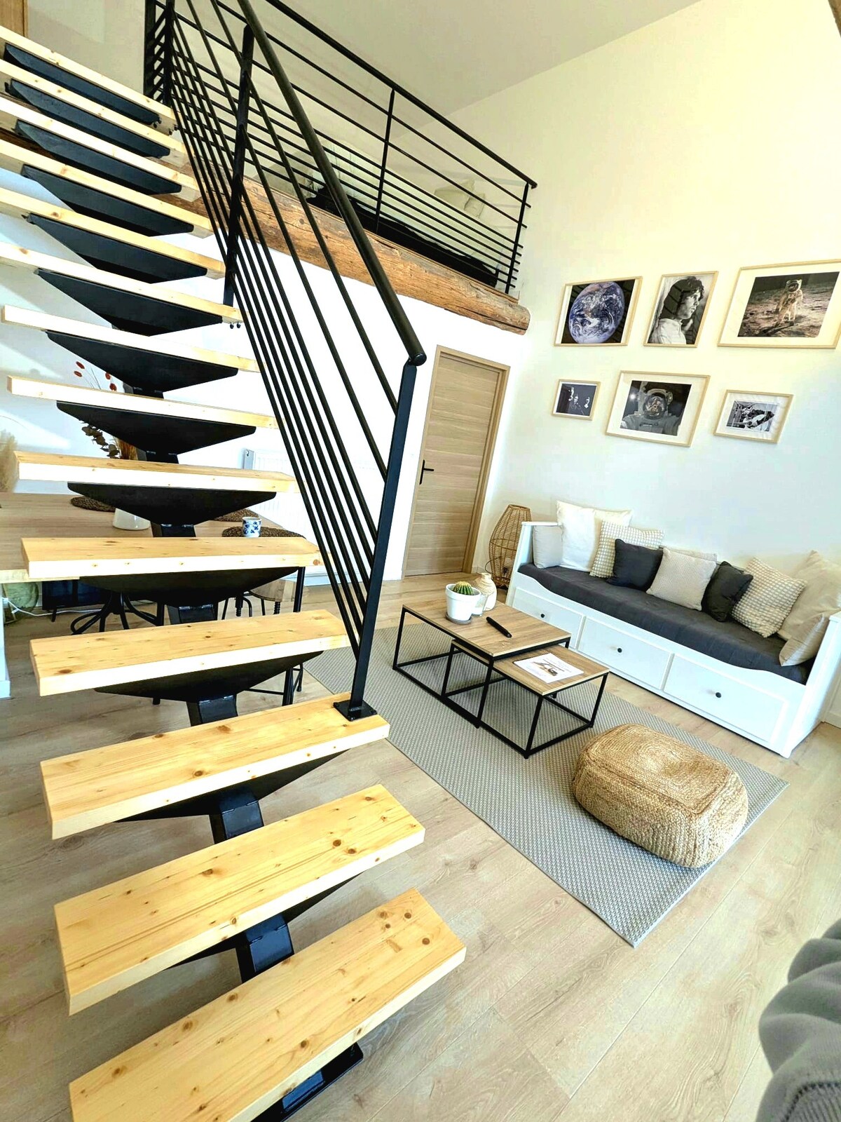 A contemporary interior features a stylish wooden staircase with black metal railings leading to a lofted sleeping area. A light-colored sofa with cushions is positioned beside a coffee table, while framed wall art adds character to the space. Natural light fills the room.