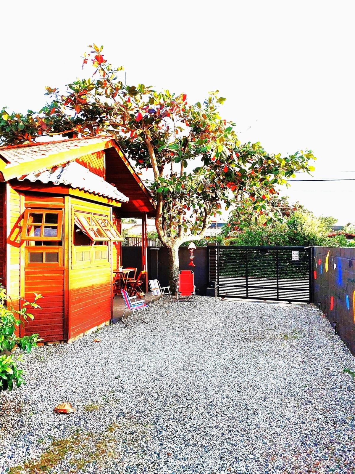 A vibrant wooden structure is framed by lush greenery and a gravel pathway. The entrance is highlighted by a welcoming porch with a few outdoor chairs. A secure, black gate stands nearby, providing access to the property. Bright sunlight casts a cheerful ambiance over the scene.