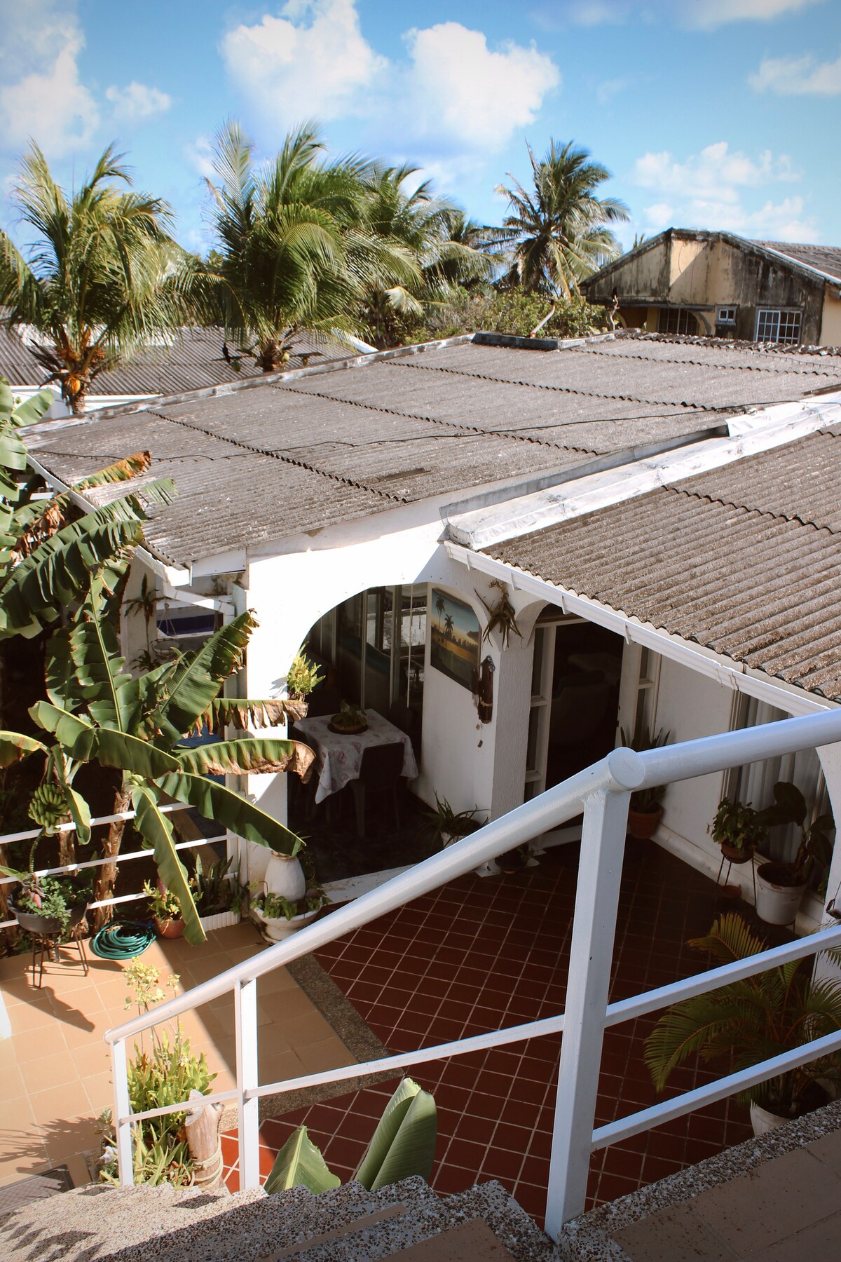 The view from above reveals a serene courtyard surrounded by lush greenery and palm trees. A white exterior structure is visible, with tiled pathways leading into the space. The overhanging roof and a staircase add depth to the tranquil atmosphere.