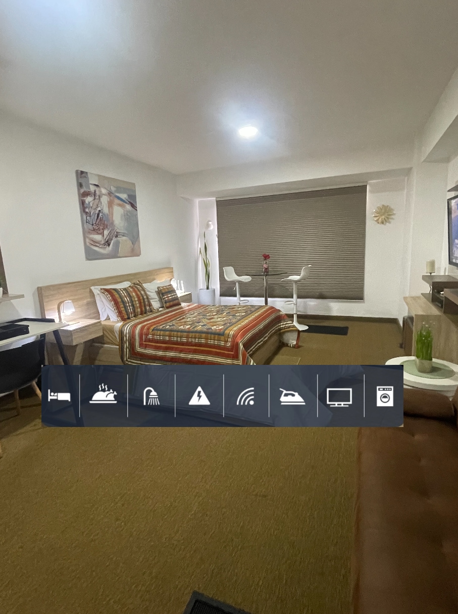 A spacious bedroom features a comfortable Queen bed dressed in a patterned blanket. A large window is covered by a roller shade, allowing for natural light. A cozy seating area with a sofa complements a glass dining table set for two. A TV is positioned on a low console.