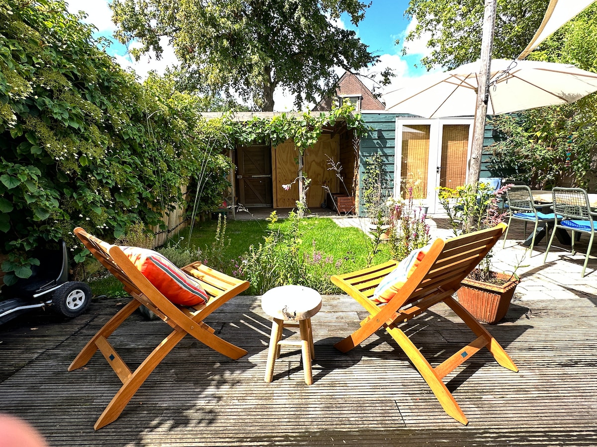 A serene garden area is presented, featuring two wooden lounge chairs with cushions and a small side table. Lush greenery surrounds the space, while a patio area offers a charming view of the neatly maintained lawn and a garden shed in the background.