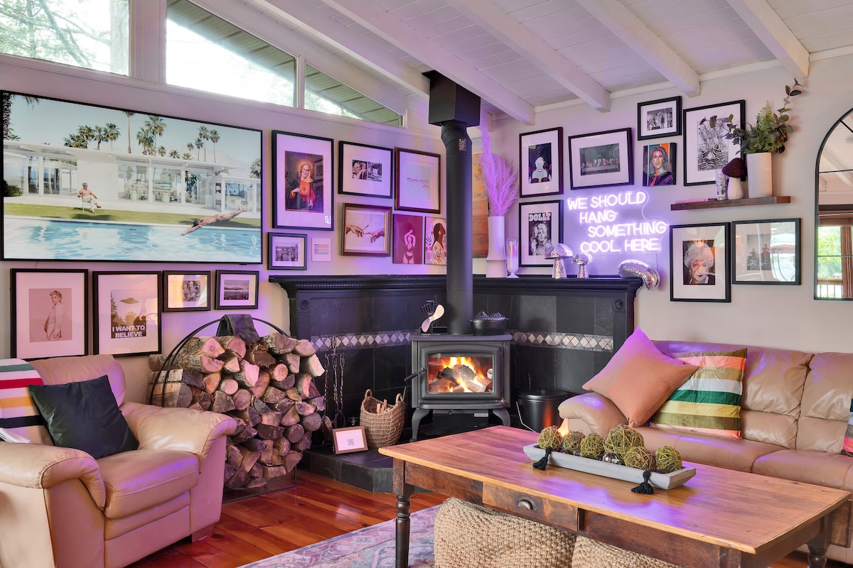 A cozy living area features a comfortable, light-colored sofa complemented by a wooden coffee table. The wall displays a variety of framed photographs, while a modern wood stove adds warmth. A stack of firewood is neatly arranged beside the stove, contributing to a homely atmosphere.