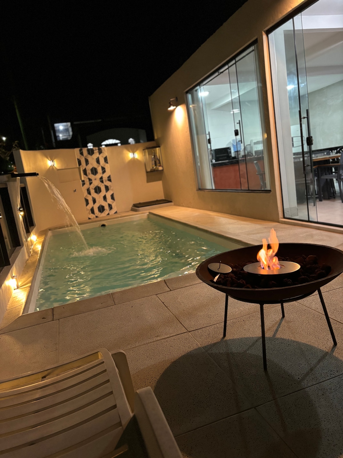 A private pool area is illuminated by soft lighting, enhancing its serene atmosphere at night. A gentle water feature flows into the pool, while a fire pit with flames flickers nearby. Large glass doors provide visibility into the interior space.