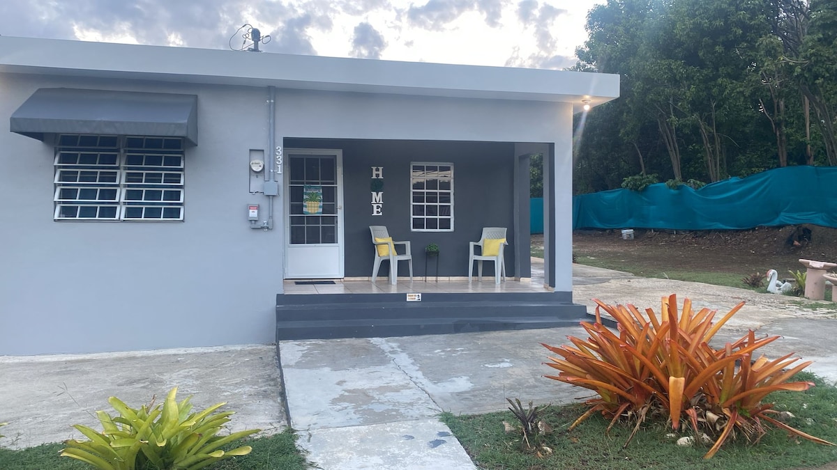 The exterior of a modern house is presented, featuring a welcoming porch with two chairs and a small potted plant. A decorative 'HOME' sign is displayed on the wall. Lush greenery surrounds the property, with a grassy area and a backdrop of vibrant plants.