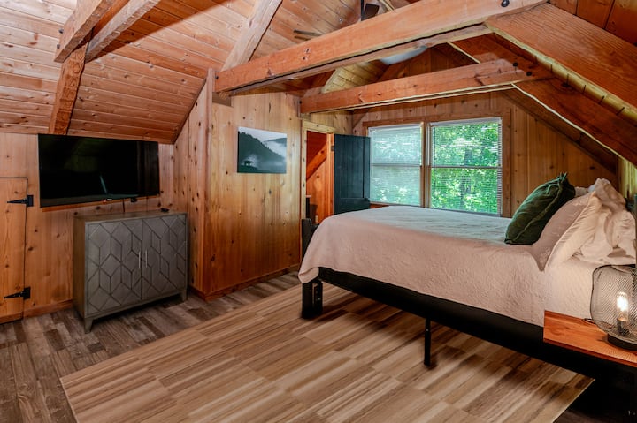 Loft bedroom tucked under sloped wood beam ceilings, with a queen log bed, wall mounted tv, ensuite half bath dresser filled with games, nightstand lamp, and a window nook.