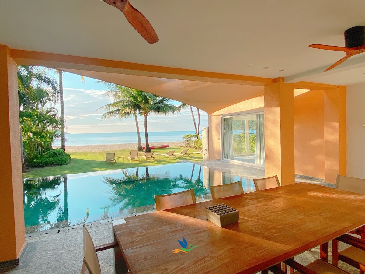 A spacious dining area is showcased with a large wooden table surrounded by several chairs. Large glass doors provide a view of the outdoor pool and beach beyond, with palm trees framing the scene under a bright sky.