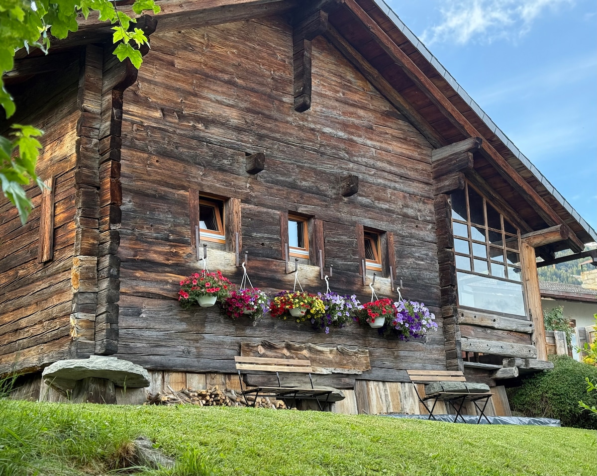 The exterior of the chalet is constructed from weathered wood and features several small windows adorned with vibrant flowerboxes. A grassy area is visible in the foreground, where two wooden tables are positioned. The building exhibits traditional alpine architecture and is set against a backdrop of clear blue skies.