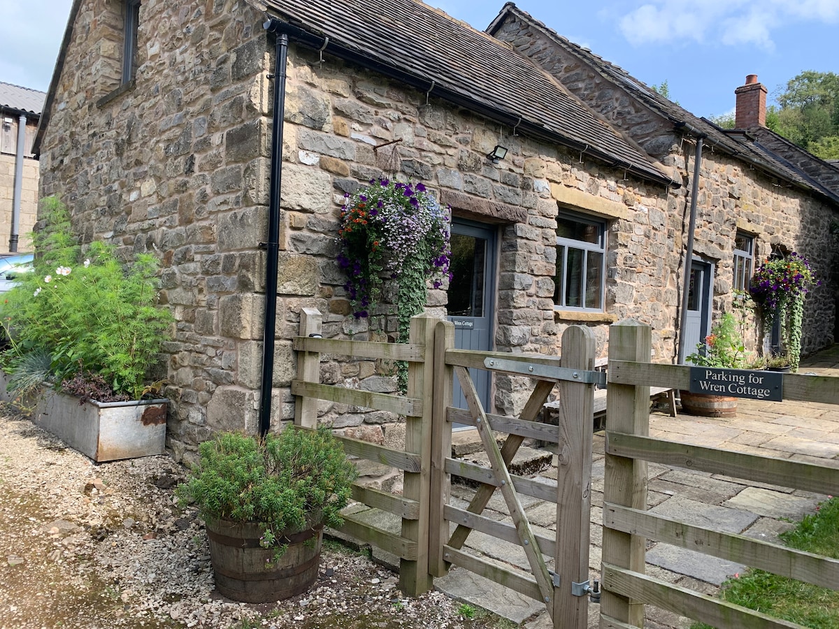 An inviting stone cottage is framed by green foliage and vibrant flower planters. A wooden gate marked 'Parking for Wren Cottage' leads to a gravel pathway. The rustic charm of the exterior is complemented by the warm tones of the stone walls, under a clear blue sky.
