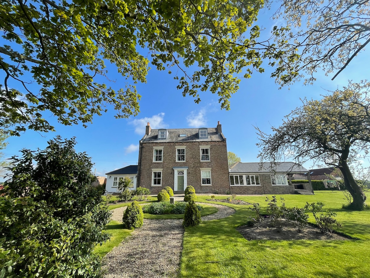 The exterior of a spacious three-story house is shown, featuring a classic brick facade. Lush greenery surrounds the property, with well-maintained gardens and a serene pathway leading to the entrance. Clear blue skies and sunlight enhance the inviting atmosphere.