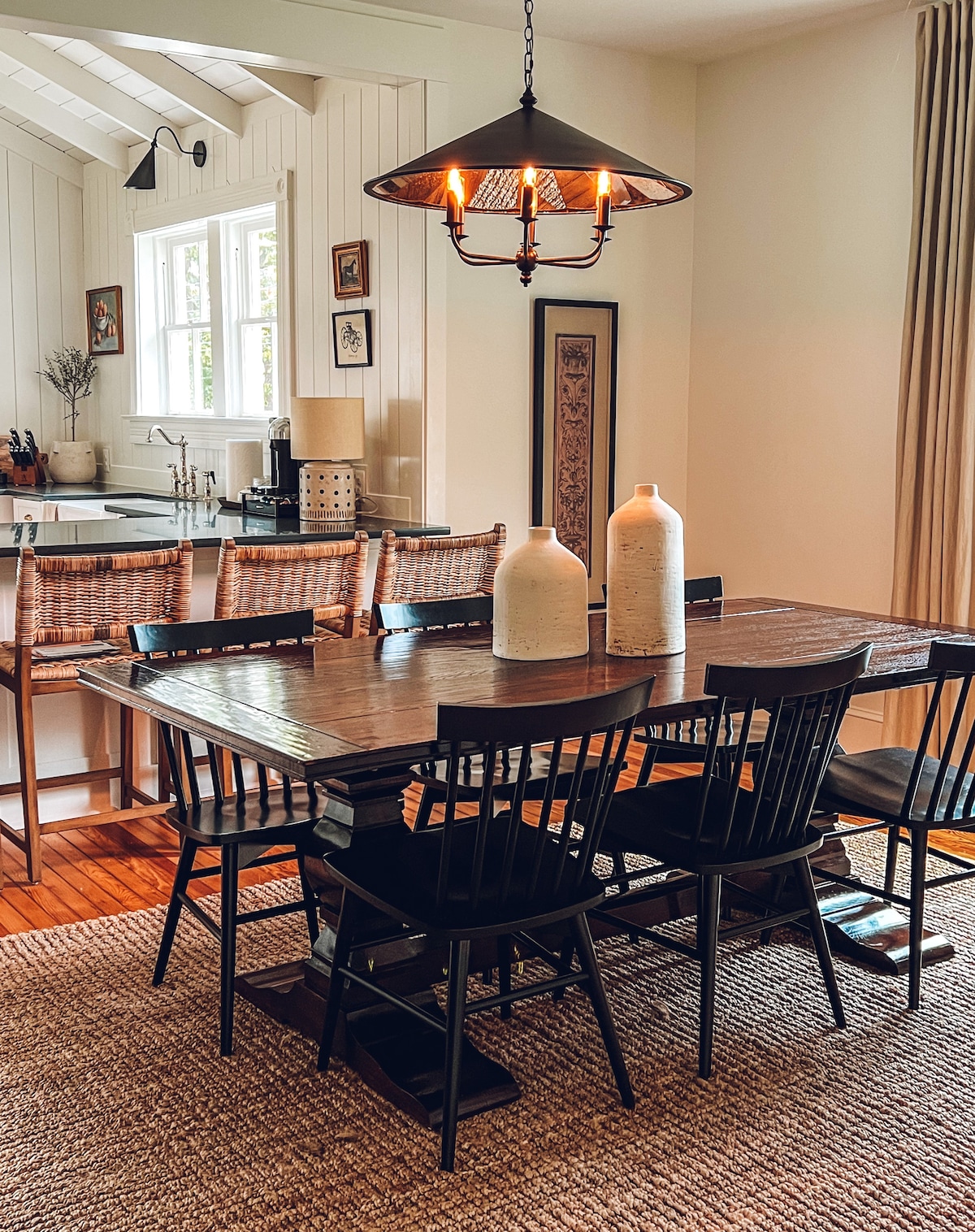 A dining area features a dark wooden table surrounded by six black chairs. Two large ceramic vases are centered on the table. A softly lit chandelier hangs above, illuminating the space, which is complemented by light-colored walls and woven textures.