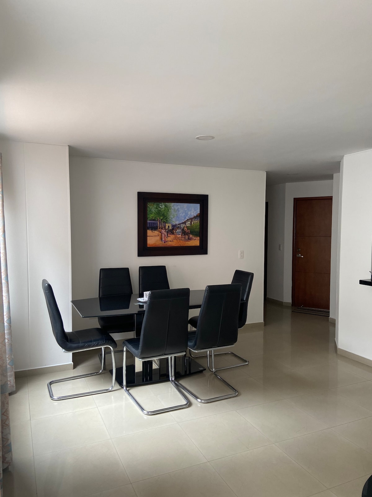 An dining area features a sleek black table surrounded by six black chairs. A framed artwork depicting a rural scene is displayed on the wall. Natural light softly illuminates the space through a nearby window.