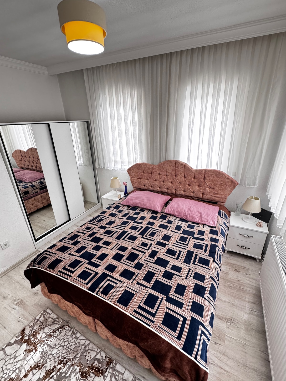 A cozy bedroom is featured with a spacious bed adorned with a patterned blanket and pink pillows. A stylish headboard complements the room, while a mirrored wardrobe reflects the soft natural light coming through the sheer white curtains. Two bedside lamps provide additional lighting.