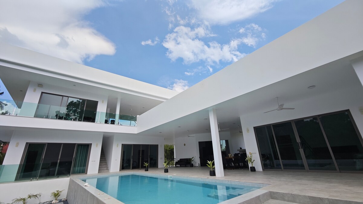 A modern villa features a spacious outdoor area, centered around a stylish swimming pool. Wide glass doors provide views into the interior spaces, while balcony railings and potted plants add to the aesthetic. The sky is partly cloudy, enhancing the brightness of the surroundings.