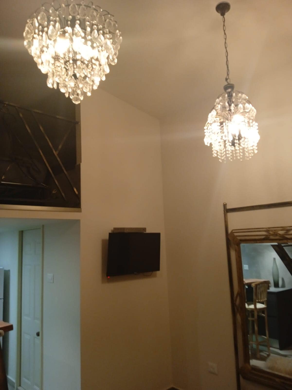 Two elegant chandeliers hang from a high ceiling, providing ambient lighting to the room. A modern flat-screen television is mounted on a neutral wall, and a large decorative mirror is positioned nearby, reflecting the softly lit space.