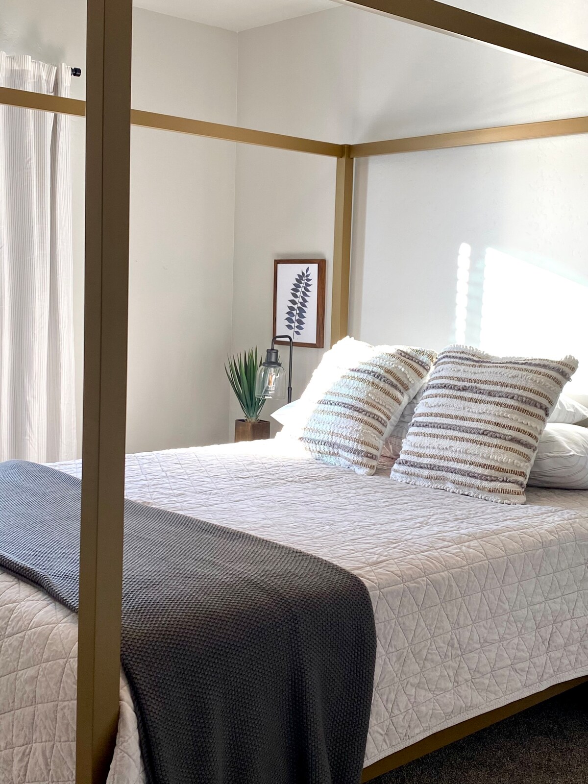 A king size bed is presented with a light quilt and decorative pillows. Natural light illuminates the space through sheer curtains, highlighting a framed botanical print and a small potted plant on a bedside table. The room features a neutral, relaxing color palette.