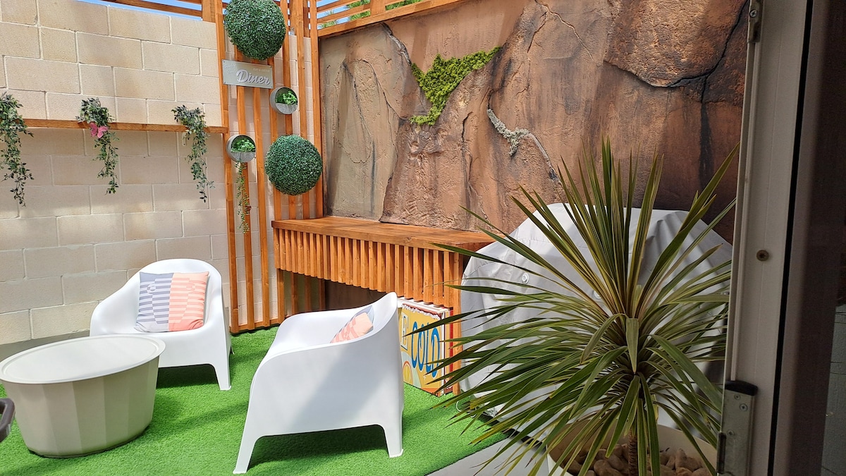 An outdoor seating area is presented with two white chairs surrounded by vibrant greenery. A stone feature serves as a backdrop, complemented by wooden accents. A circular coffee table sits centrally, and a tropical plant adds a touch of nature to the tranquil space.