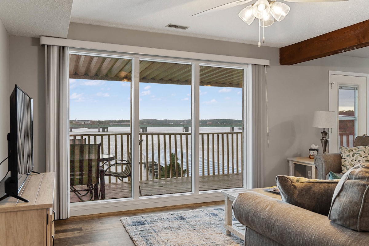 A living room area features a large sliding glass door that opens to a private deck overlooking the water. Comfortable seating arrangements are provided along with a flat-screen TV. Natural light enhances the space, creating a welcoming atmosphere.