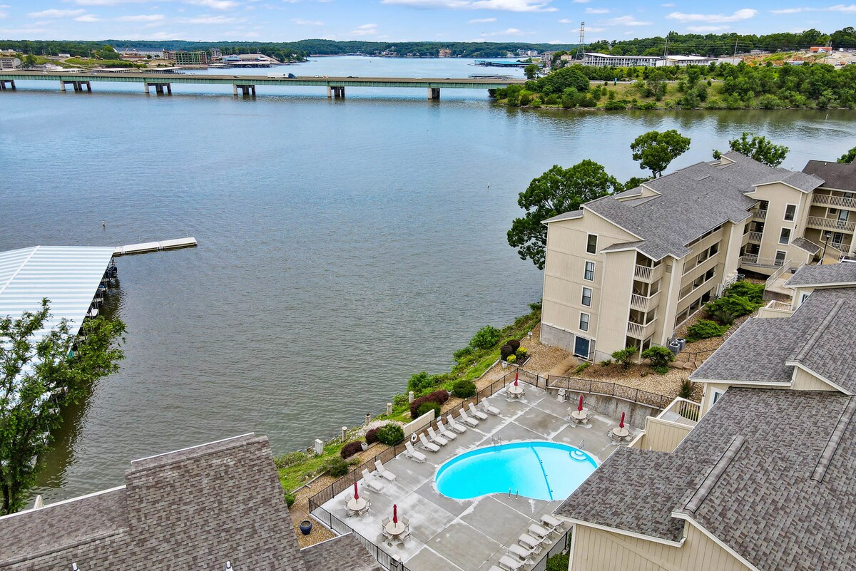 The image captures a scenic view of a serene lake, with a swimming pool situated near the water's edge. Surrounding green trees frame the area, while a bridge spans across the lake in the background, enhancing the tranquil atmosphere of the location.