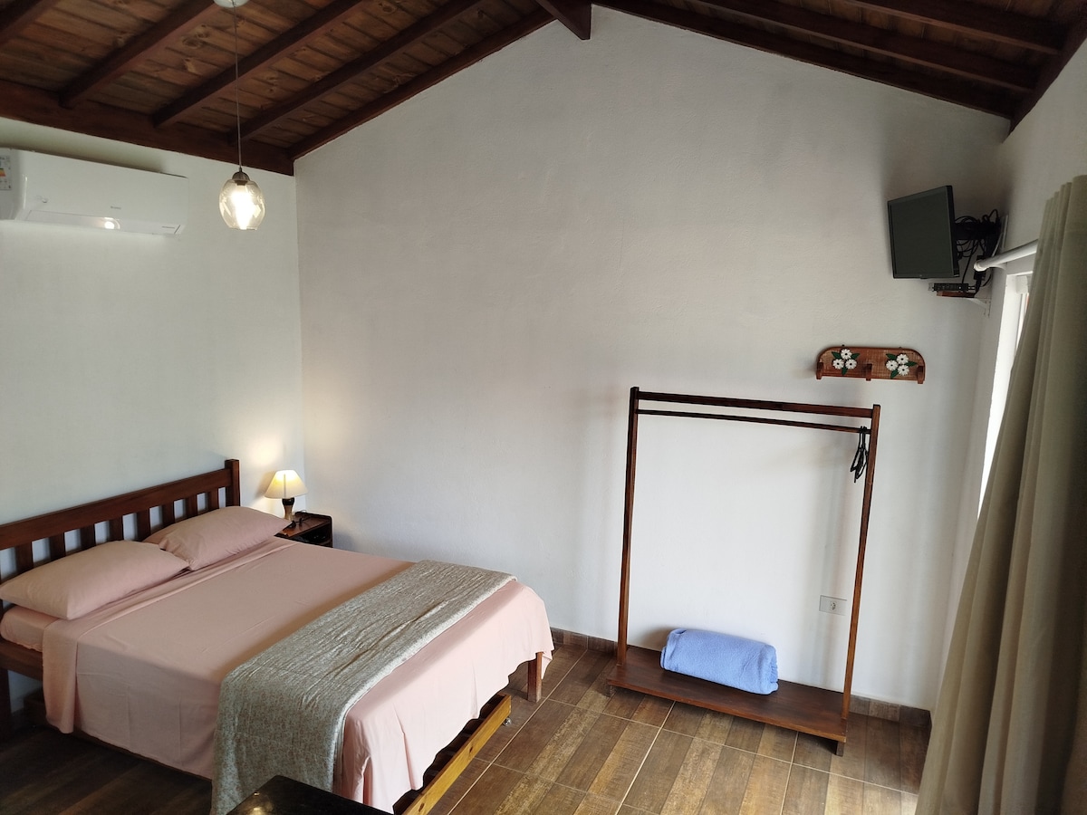 A spacious sleeping area is featured, containing a bed with light bedding and a wooden frame. A simple clothing rack stands nearby, while a small television is mounted on the wall. Natural light enters through a window with sheer curtains.