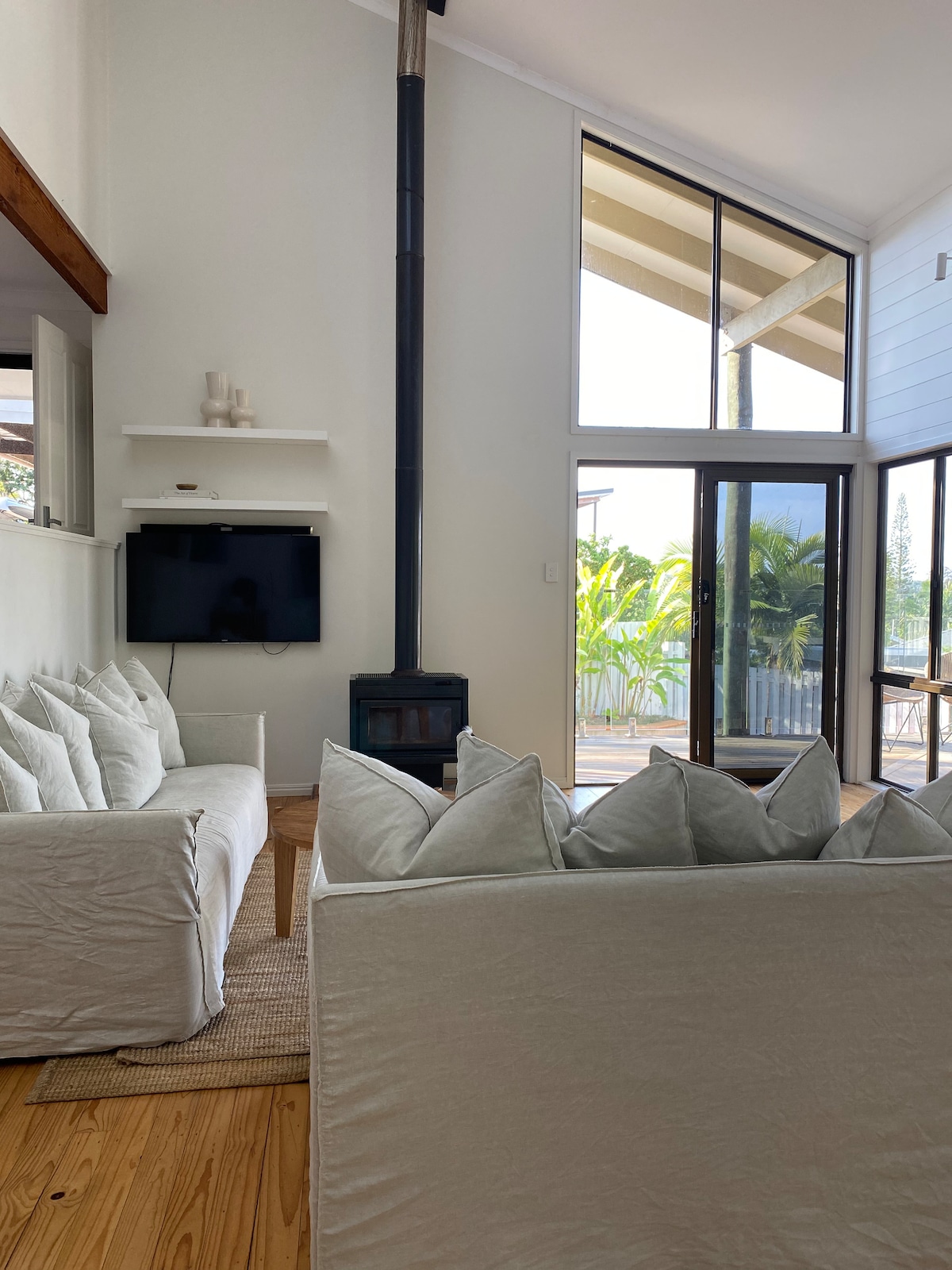 Two light-colored, comfortable sofas are positioned around a natural fiber rug in a bright living area. A wood stove is mounted along the wall, with large windows allowing natural light and views of greenery outside.