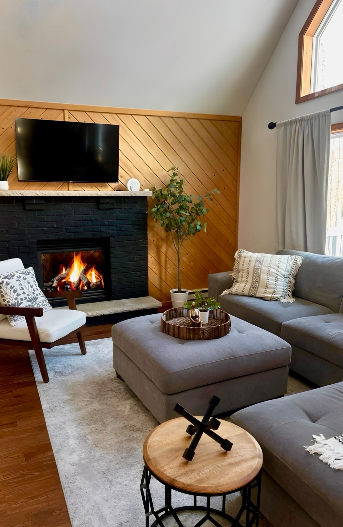 A cozy living area features a black stone fireplace, complemented by a television mounted above. Gray sectional seating surrounds a circular coffee table with decorative items. Natural light enters through a large window, illuminating the space with a warm ambiance.