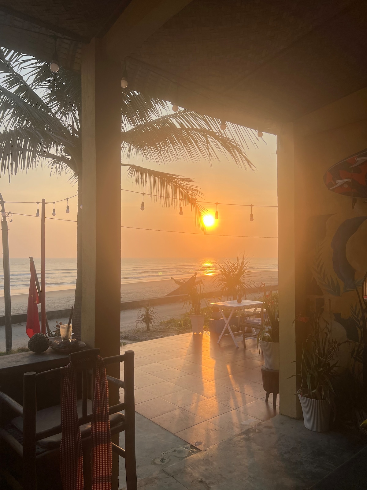 A serene beach view is presented, with soft golden sunlight reflecting off the ocean waves. The patio area is adorned with potted plants, and decorative string lights are suspended overhead, enhancing the tranquil ambiance of the sunset scene.