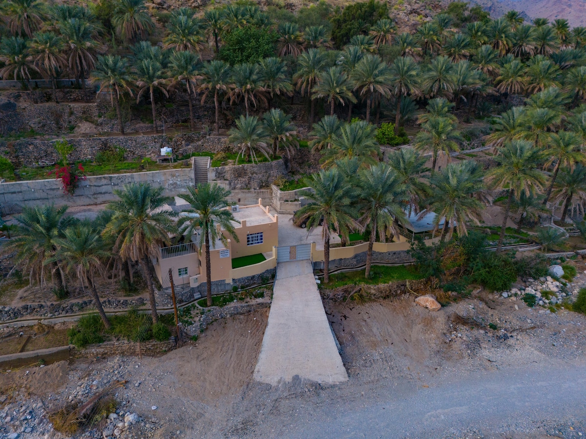 The garden house is nestled among lush palm trees, with a pathway leading to its entrance. The structure features a light-colored exterior, blending with the natural surroundings. Nearby rocky terrain and greenery complement the tranquil landscape, highlighting the serene environment.