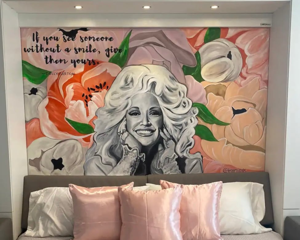 A wall mural features a design adorned with large, colorful flowers, with a quote that encourages sharing smiles. A comfortable bed is positioned below, layered with plush pillows in soft pink hues, creating a cozy and inviting sleeping area.