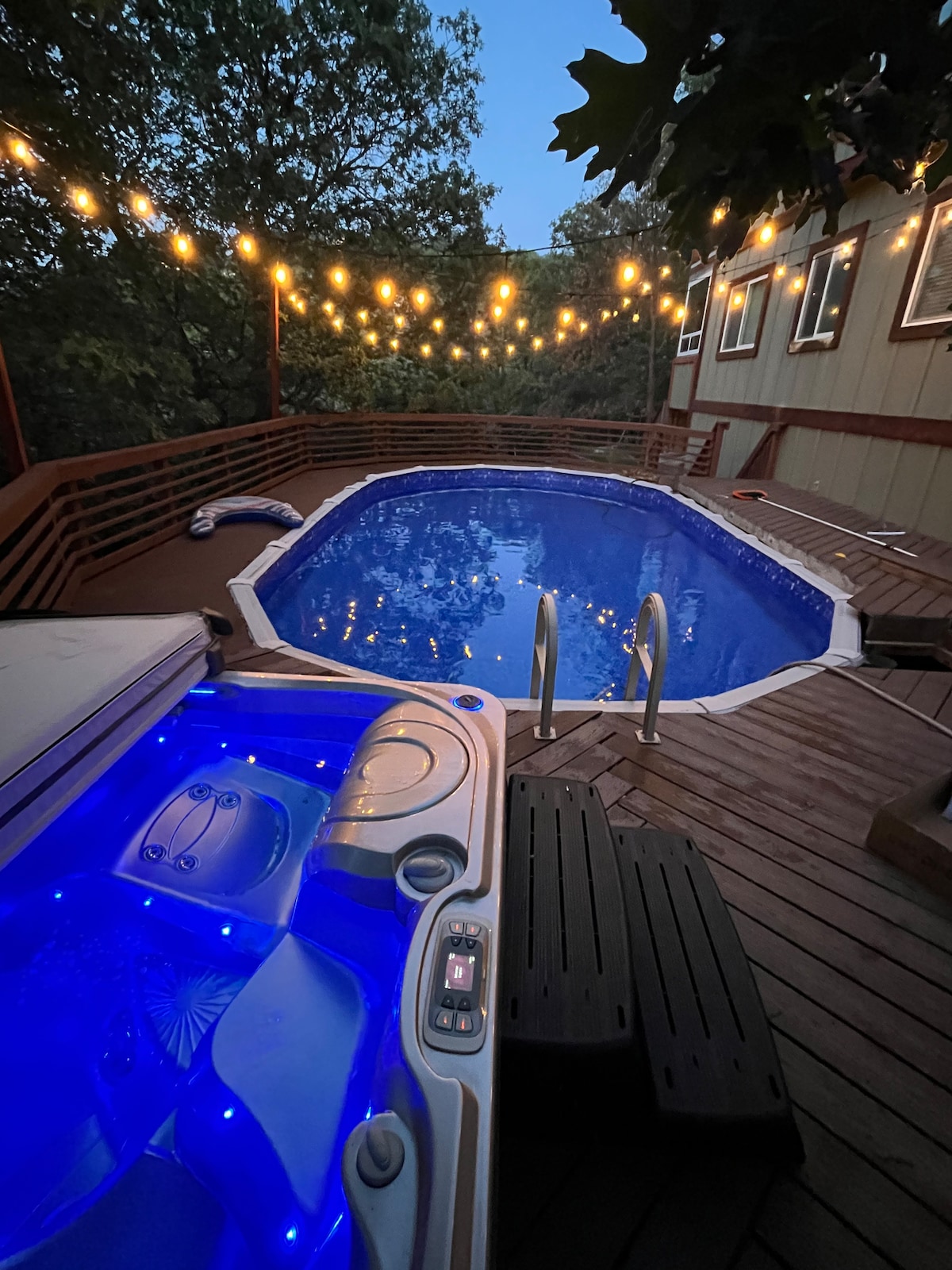 A spacious wooden deck features a well-lit private pool and a jacuzzi, with vibrant blue lights highlighting the water. String lights overhead create a cozy ambiance against the backdrop of lush greenery. The pool is easily accessible with steps leading into it.