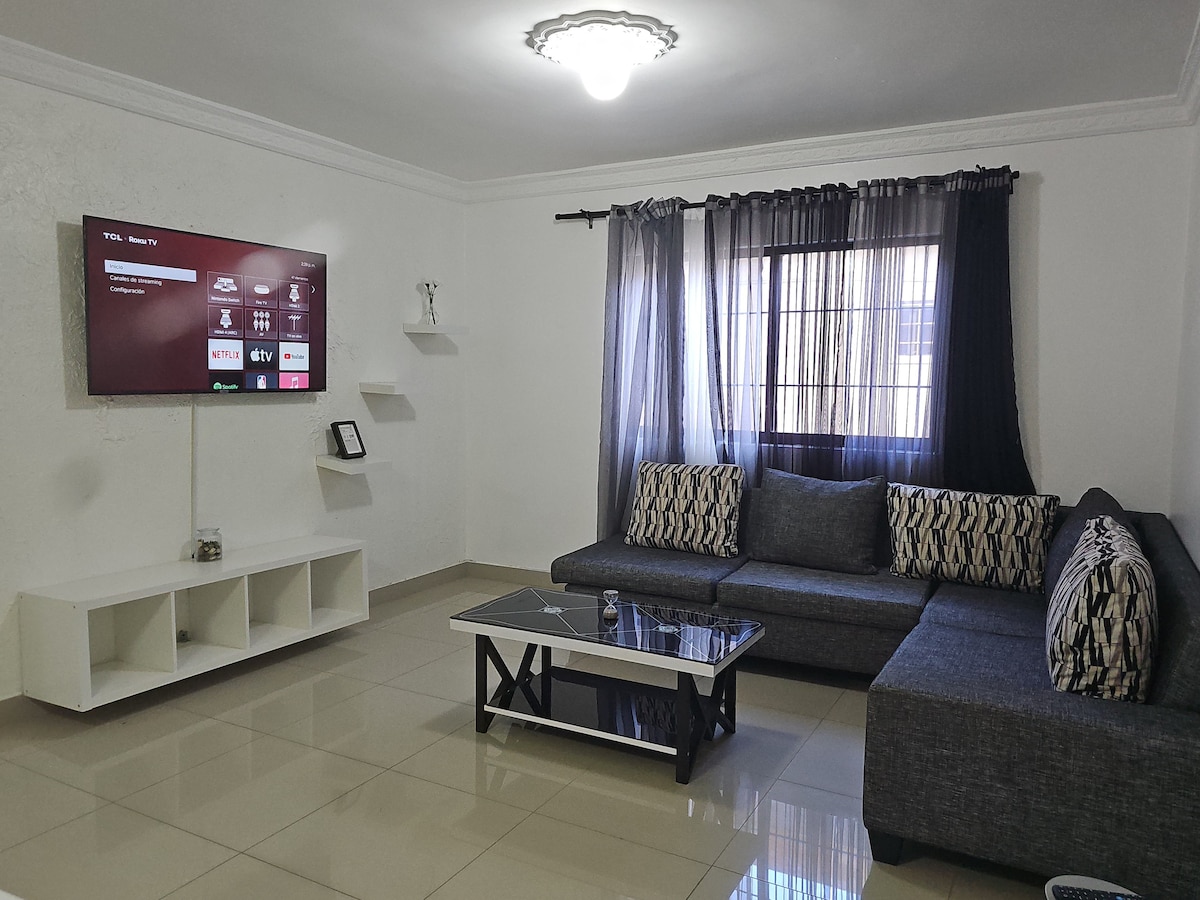A modern living area features a sectional sofa with contrasting cushions, arranged around a sleek coffee table. An entertainment unit holds a flat-screen television. Natural light filters through a window, partially covered by sheer curtains, enhancing the space's brightness.