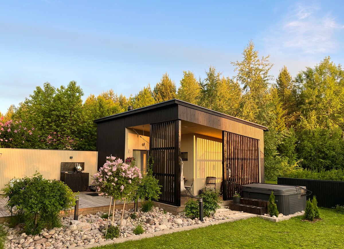 An exterior view of a separate structure surrounded by greenery. The building features a modern design with wooden slats, large windows, and a spacious deck. A jacuzzi is located on the patio, complemented by lush plants and a gravel pathway.