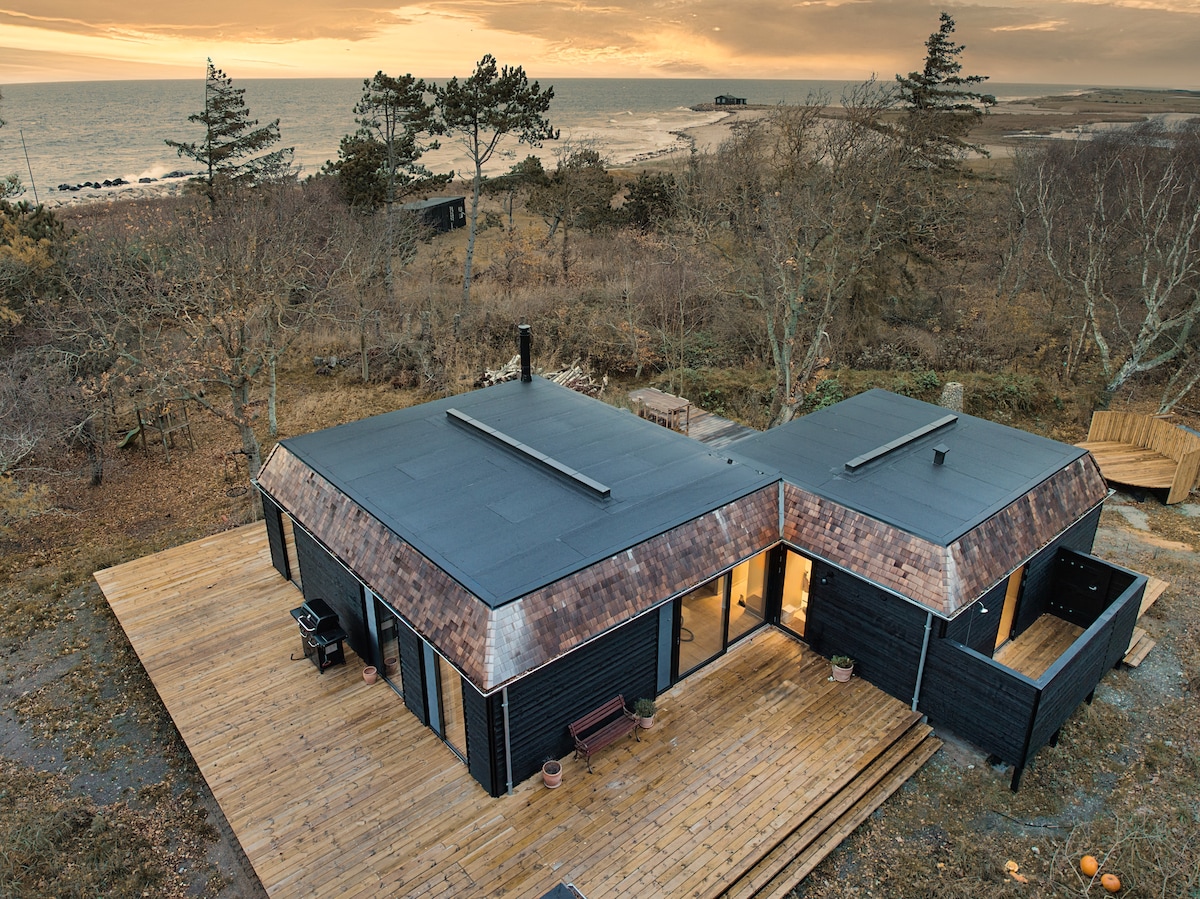An aerial view of a modern summer house with a dark facade and metal roof. Surrounding it is a spacious wooden deck that integrates with the natural landscape. The ocean can be seen in the background, framed by sparse trees under a moody sky.