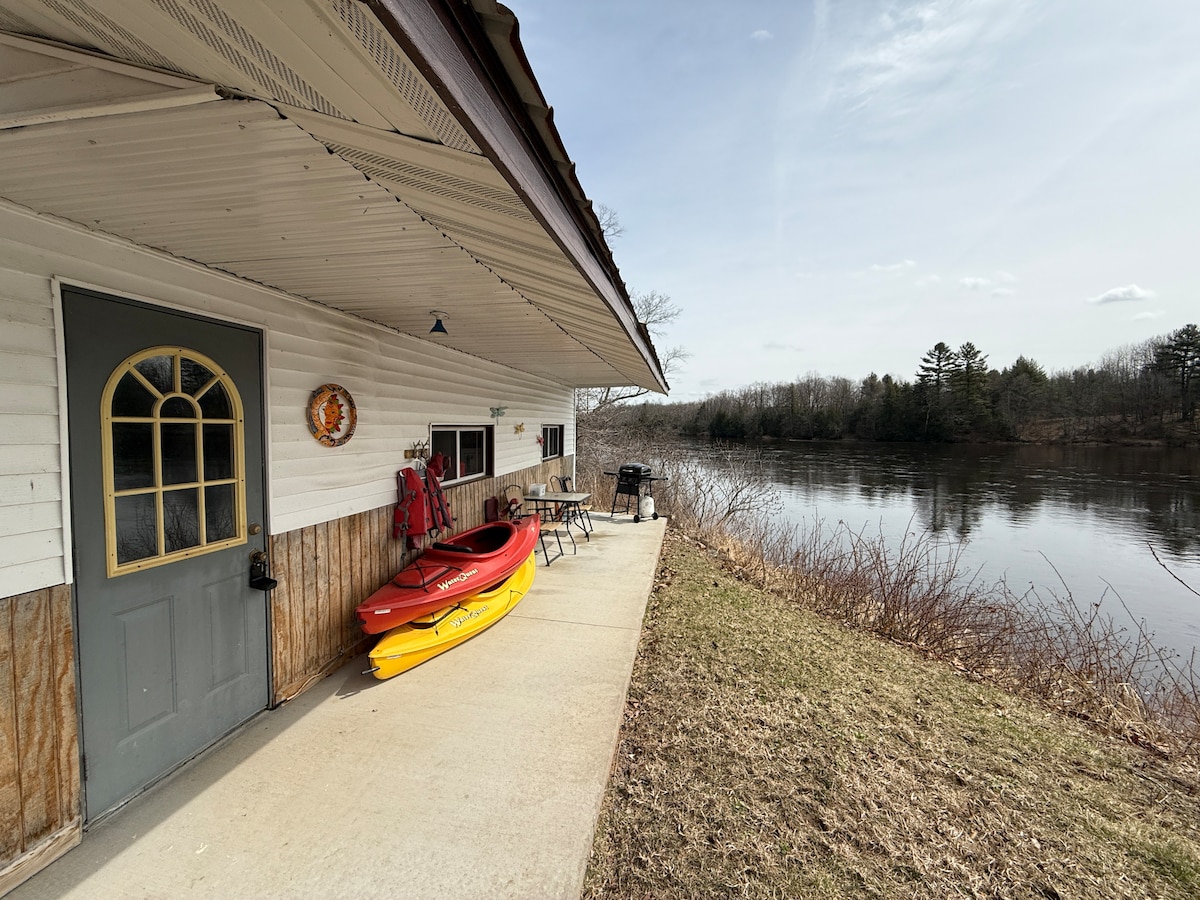 A covered patio extends along the riverbank, featuring two bright yellow kayaks. A grill and outdoor table are positioned nearby, providing a space for relaxation. The peaceful river reflects the surrounding trees and sky, creating a serene outdoor atmosphere.