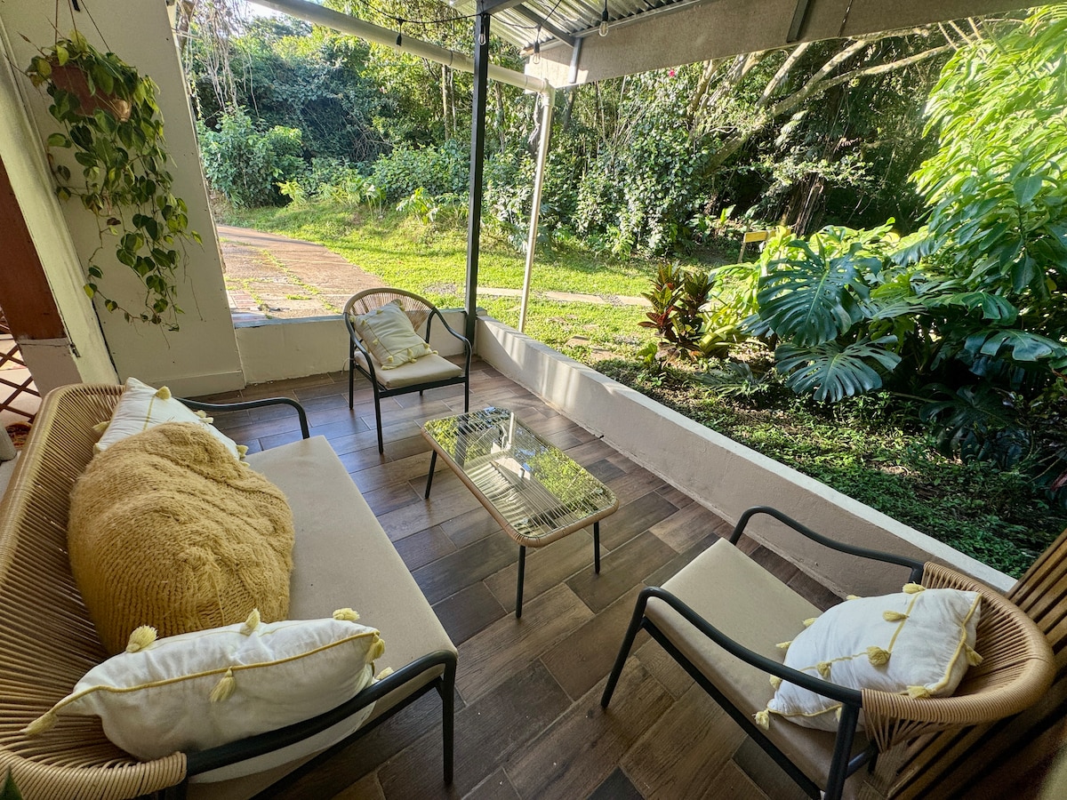 A covered terrace features two comfortable chairs and a small coffee table, surrounded by lush greenery. Soft cushions add a touch of warmth. Sunlight filters through the canopy, illuminating the natural beauty of the surrounding landscape.