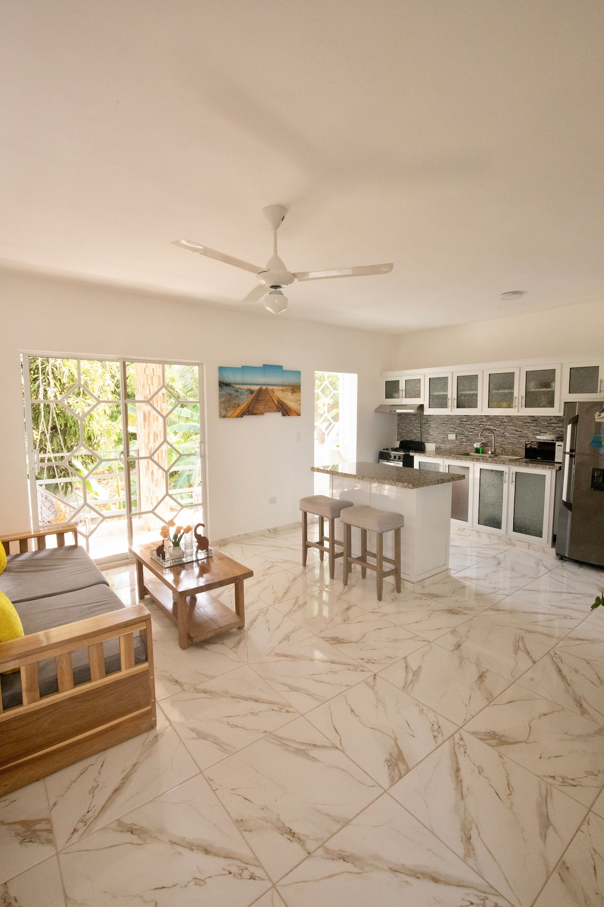 A bright living space is filled with natural light, featuring a wooden sofa and a coffee table. The open kitchen showcases modern appliances and counter space, all set against a backdrop of tiled flooring. Large glass doors open to a lush outdoor area.