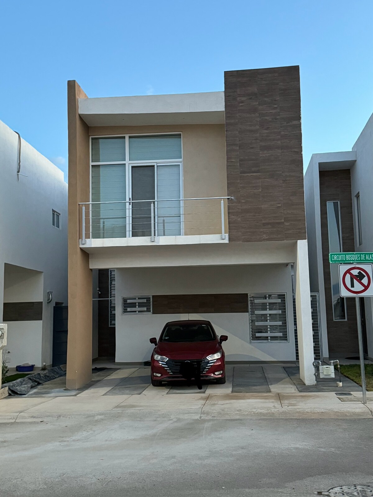 A modern two-story house is depicted, featuring a beige and brown facade. The entrance is framed by large windows, and a balcony extends from the upper level. A compact car is parked in a designated area in front, with smooth pavement leading up to the house.