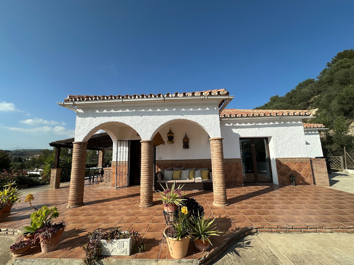 The villa features a charming entrance with arched columns and terracotta tiles. Potted plants are arranged around the outdoor seating area. The expansive terrace allows for outdoor relaxation, with views of the surrounding landscape evident in the background.