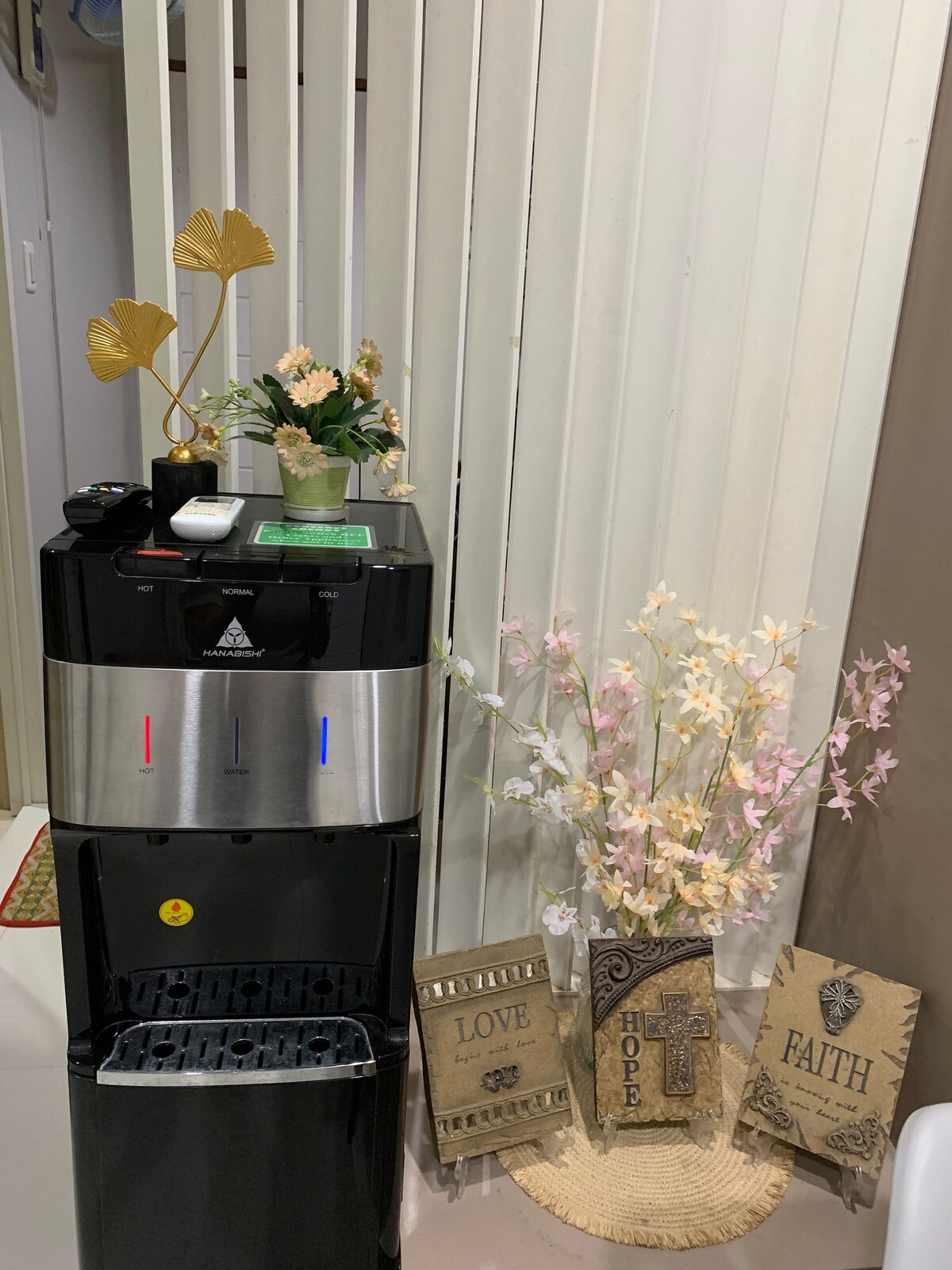 A water dispenser stands against a wall, featuring a sleek black design with silver accents. Potted plants adorn the top, accompanied by decorative signs that read 'LOVE,' 'HOPE,' and 'FAITH.' Soft pastel flowers add a touch of color beside the dispenser.