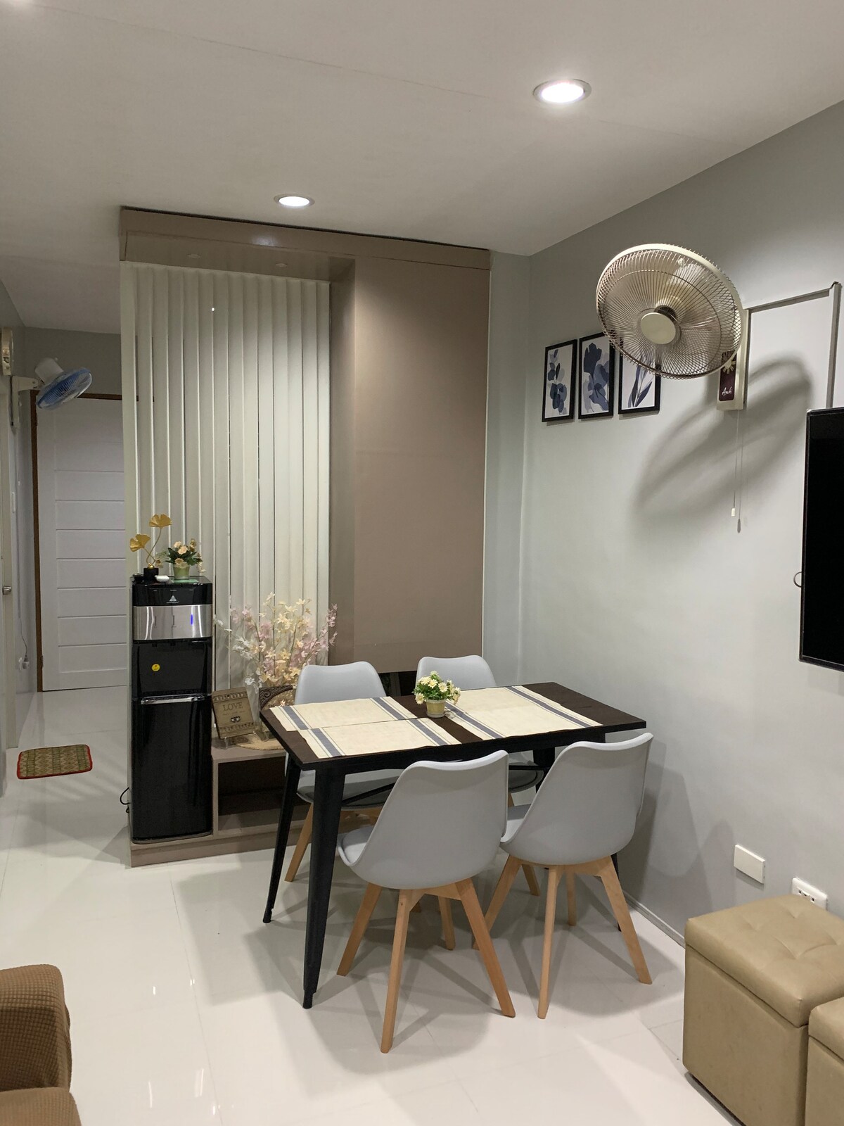 A small dining area features a dark table surrounded by four light-colored chairs. A water dispenser stands beside the table, and a floral arrangement adds a touch of decor. The wall is adorned with framed photos, and a fan is mounted for air circulation.