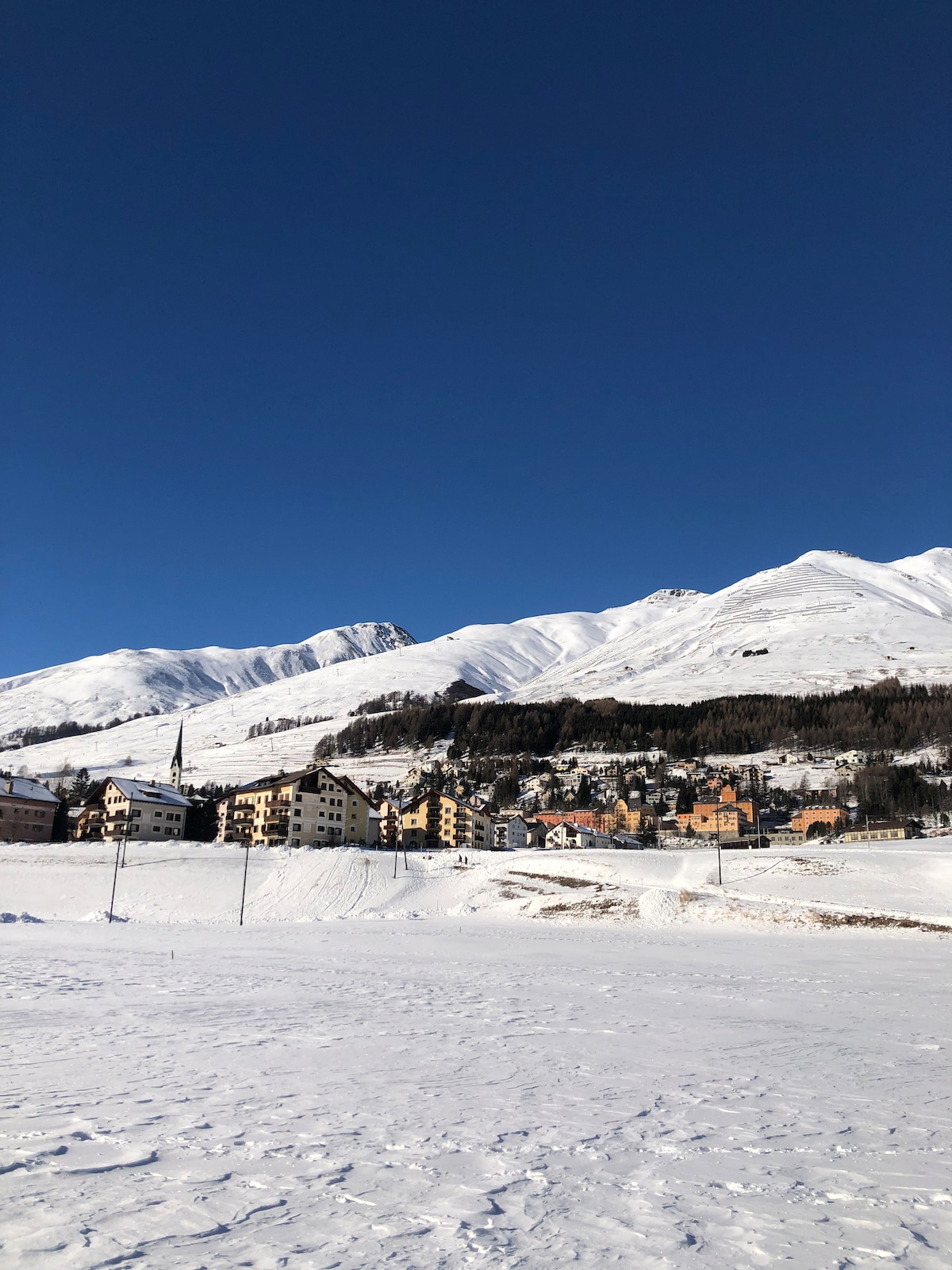A snowy landscape features a cluster of buildings nestled at the base of majestic mountains. The sky is clear and blue, providing a stark contrast to the pristine white snow covering the ground and slopes.