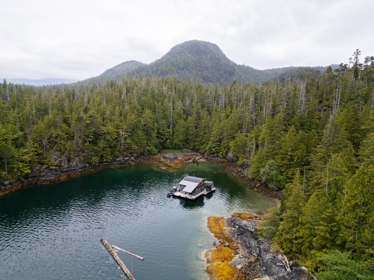 An aerial view captures the float house situated in a secluded cove, surrounded by towering trees and lush greenery. The tranquil, clear waters reflect the natural beauty of the landscape, while rocky outcrops and patches of orange seaweed add color to the shoreline.