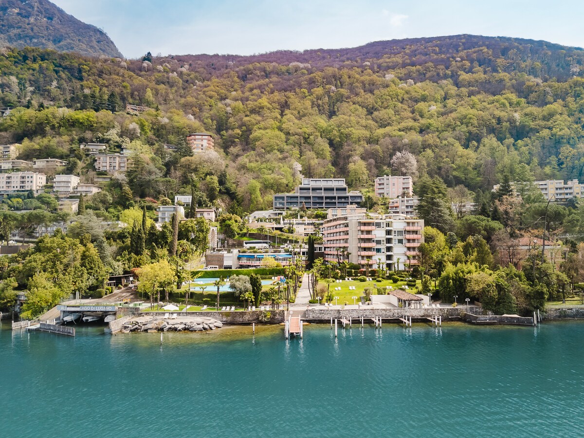 The image reveals a tranquil lakeside scene with a lush green hillside providing a backdrop. Several buildings are visible, situated alongside a well-maintained lawn leading to the water's edge. The clear blue waters reflect the surrounding greenery, contributing to a serene atmosphere.