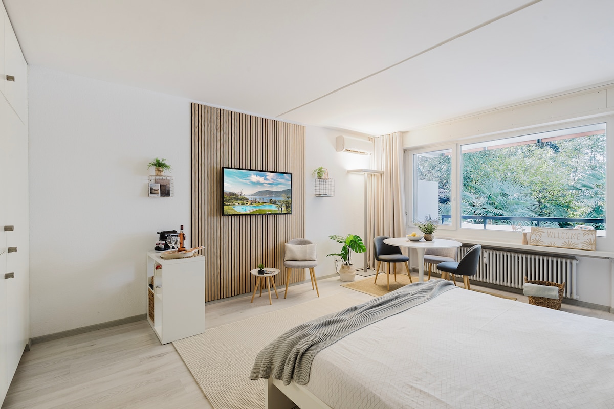 The living and sleeping area features a spacious bed with a light-colored blanket. A flat-screen TV is mounted on the wall, complemented by neutral wood paneling. A window offers views of greenery, while a small dining nook with two chairs is positioned beside it.