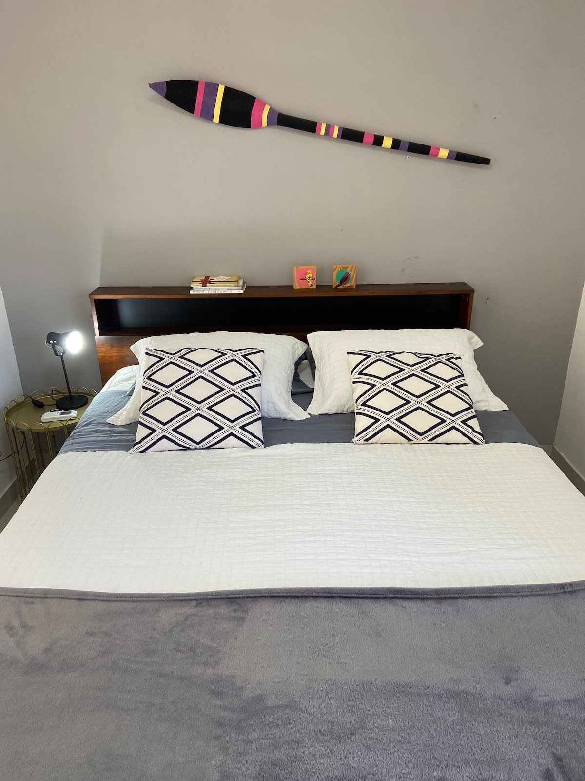 A cozy sleeping area features a spacious bed dressed with white and gray bedding. Two decorative pillows are placed on each side of the bed. A bedside table with a reading light stands next to the bed, while a large decorative paddle hangs above the headboard.