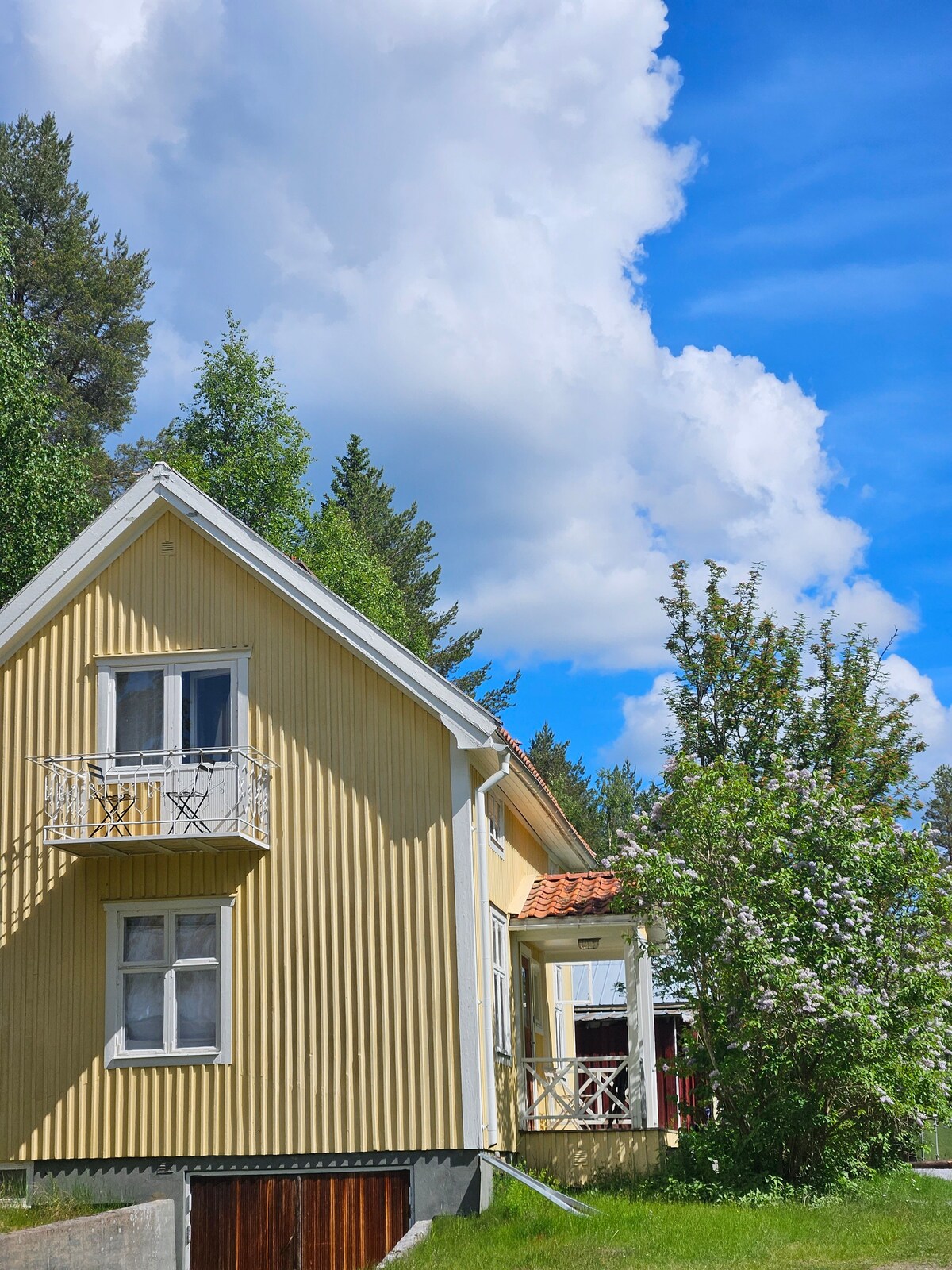 A charming yellow house stands against a vibrant blue sky adorned with fluffy clouds. The structure features a prominent balcony with decorative railings, while lush greenery surrounds the property, highlighting a serene rural setting.